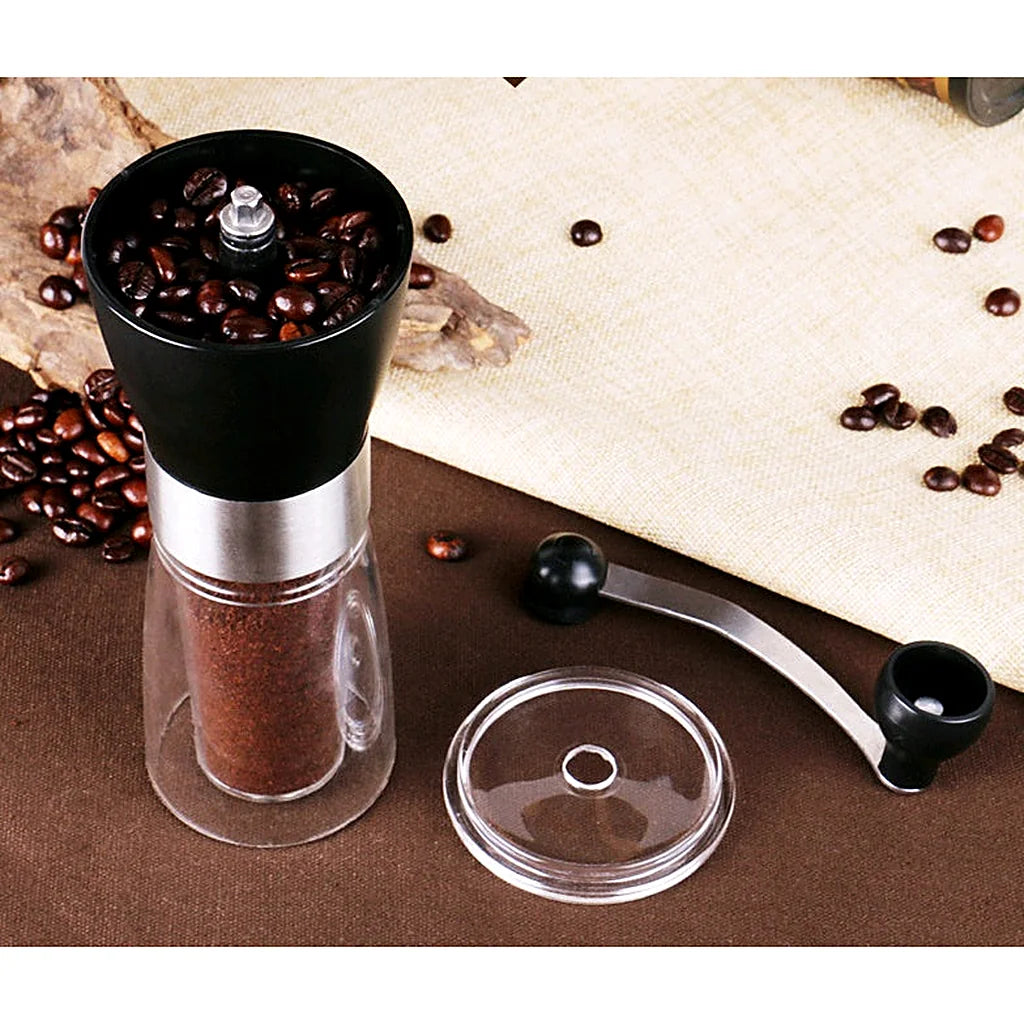 Open manual coffee grinder filled with whole beans and ground coffee, with its crank handle and clear lid beside it