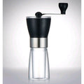 Assembled black hand-crank coffee grinder with clear glass collection jar against a soft grey studio background