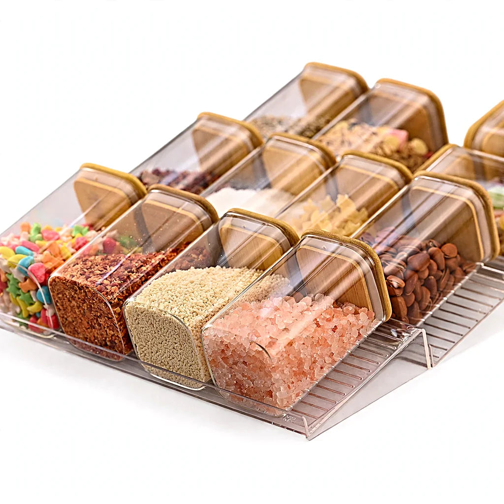 Tiered clear spice rack holding tilted jars with bamboo lids filled with pink salt, sesame, chili flakes and beans