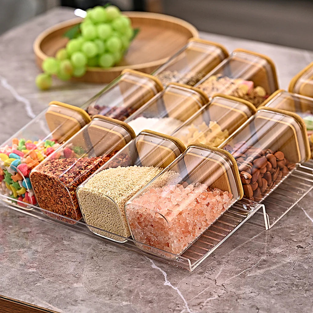 Tiered spice rack with filled jars on a grey marble countertop, wooden tray of green grapes behind it