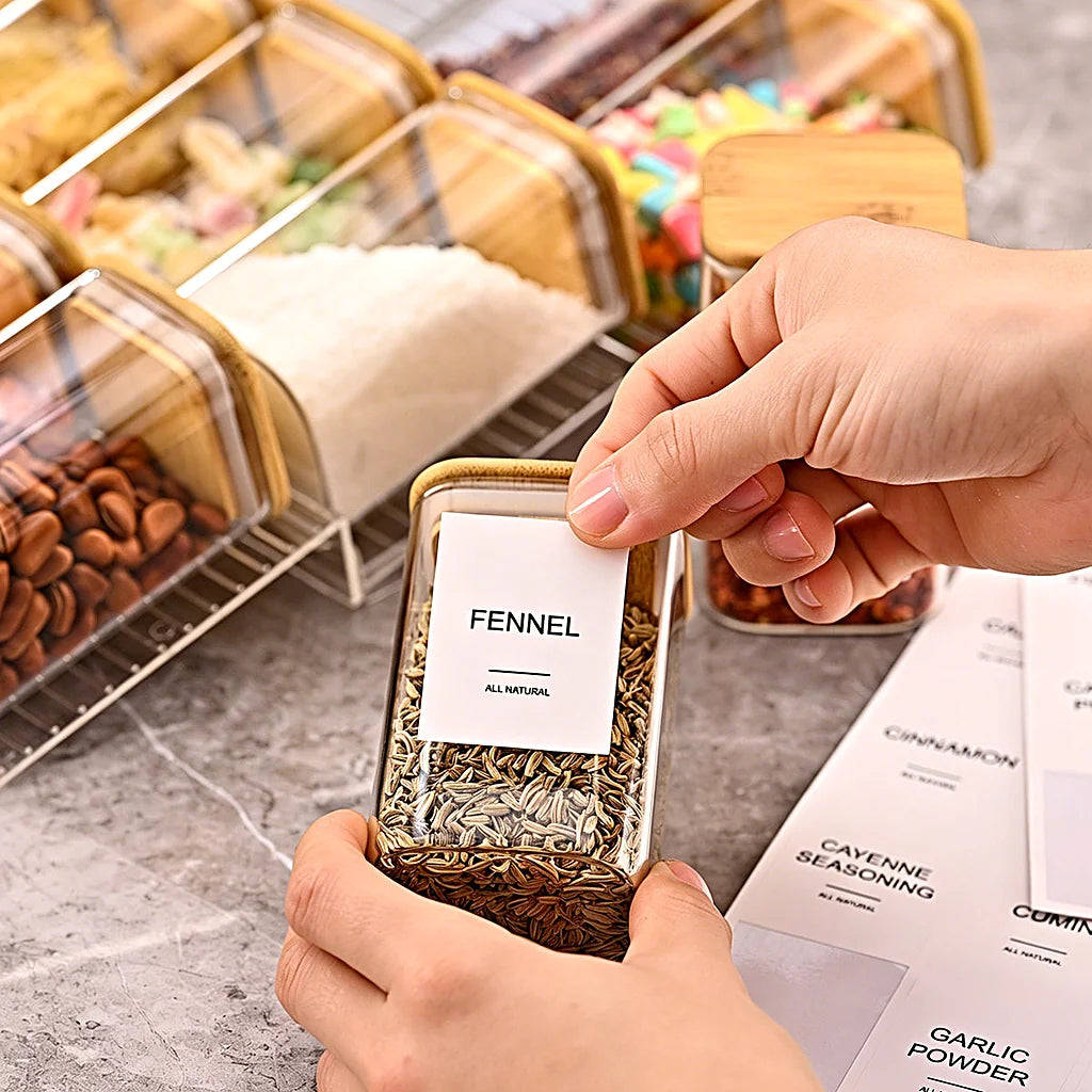 Hands applying a fennel label to a clear spice jar, with a sheet of spice labels and the filled rack nearby