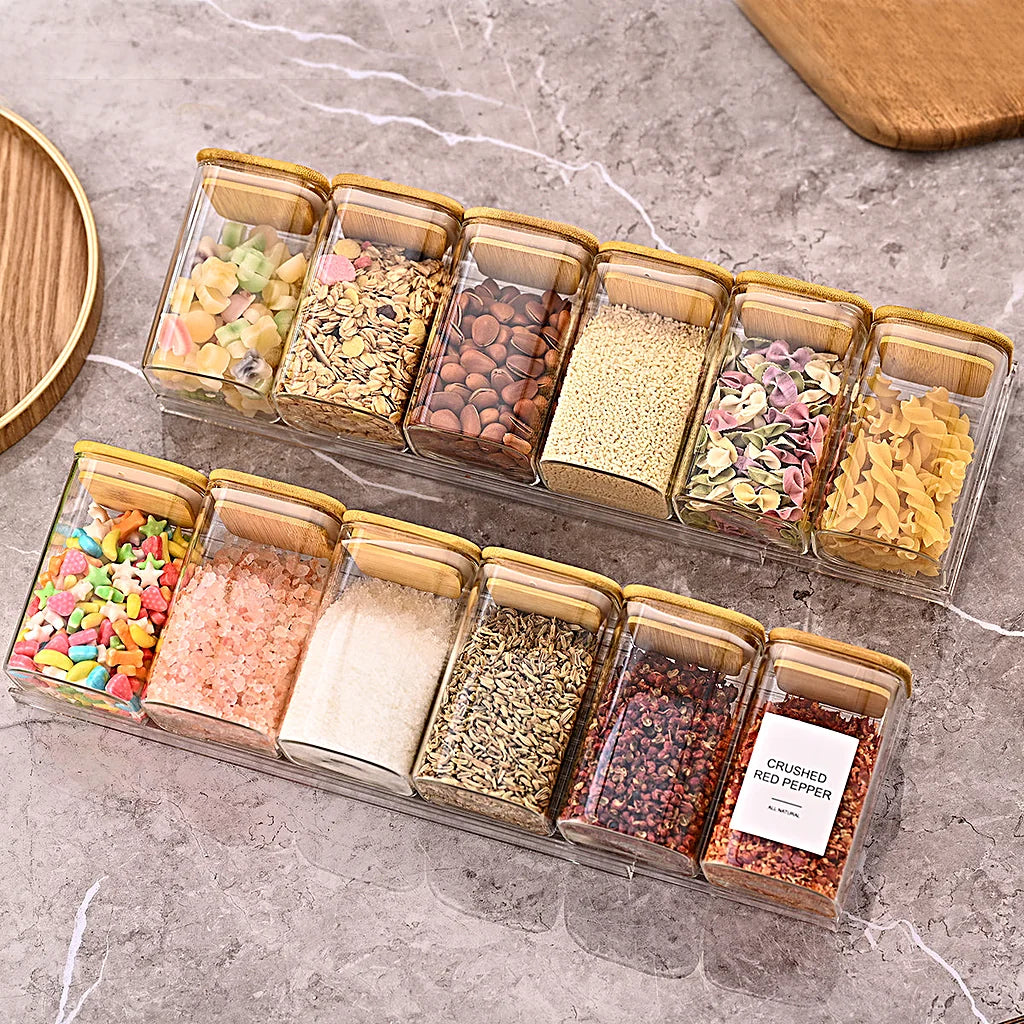 Two rows of clear spice jars with bamboo lids in clear trays on a grey marble countertop