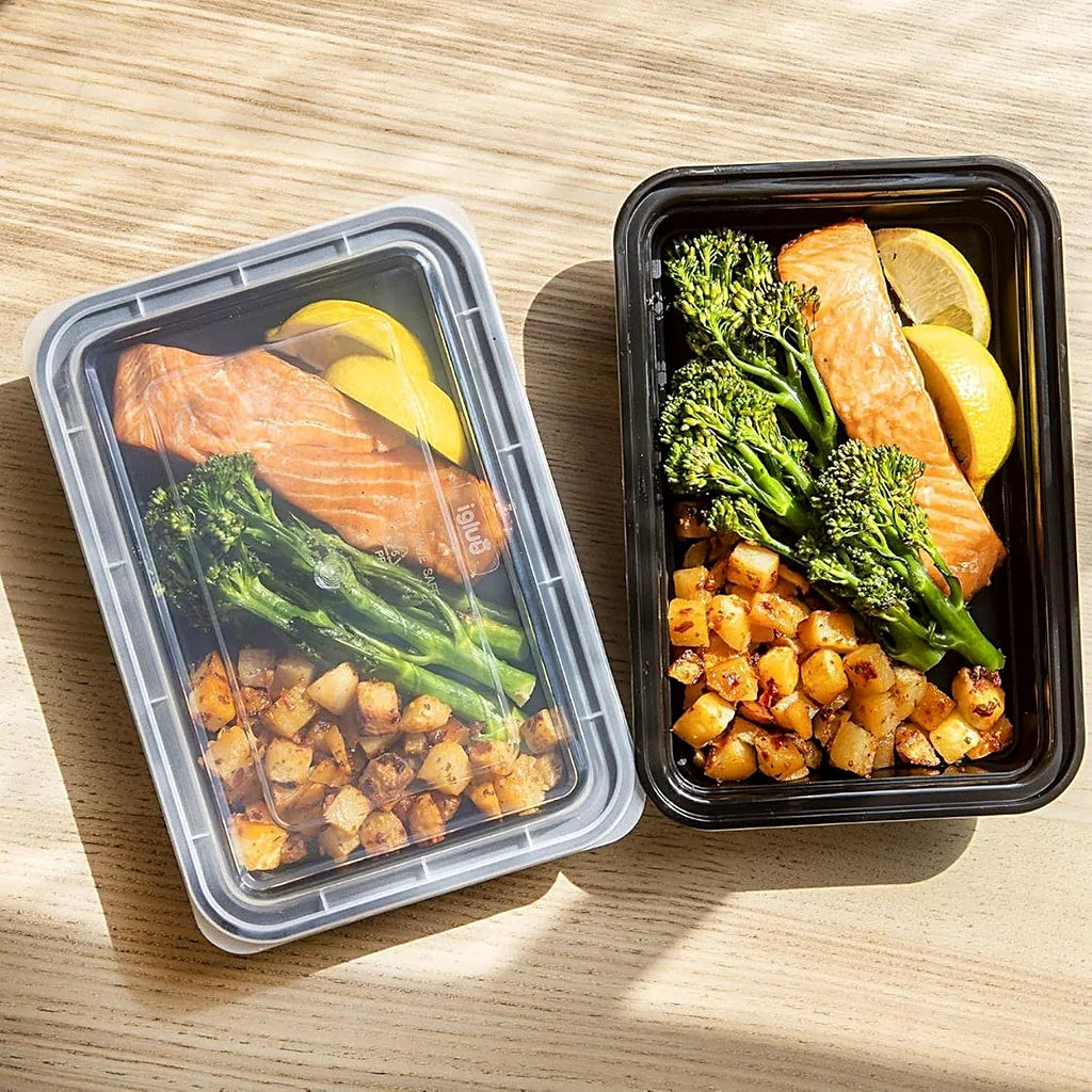 Two black meal prep containers on a wooden table with salmon, broccolini, diced potatoes and lemon, one with a clear lid on