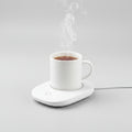 A white electric mug warmer holding a white ceramic mug filled with tea, with steam rising, on a light gray background.