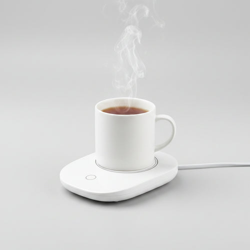 A white electric mug warmer holding a white ceramic mug filled with tea, with steam rising, on a light gray background.